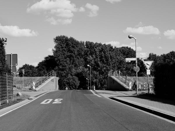 Week 26 - Dike Gate. The dike gate in Langst-Kierst is the only breach in the Rhine dike on Meerbusch territory. The dike gate was completely rebuilt in the course of the dike renovation in 2010 and considerably widened in the process. Whereas previously only one car at a time could pass through the culvert, since then there have been two lanes and a wide pedestrian and bicycle path. In case of imminent flooding, the gate can be closed with two rows of aluminum beams. However, this has so far only been done for practice purposes with the new dike gate. The road ends after a short distance at the ferry terminal of the Rhine ferry to Düsseldorf-Kaiserswerth. Immediately behind the trees in the picture, the Rhine bank has already been reached.